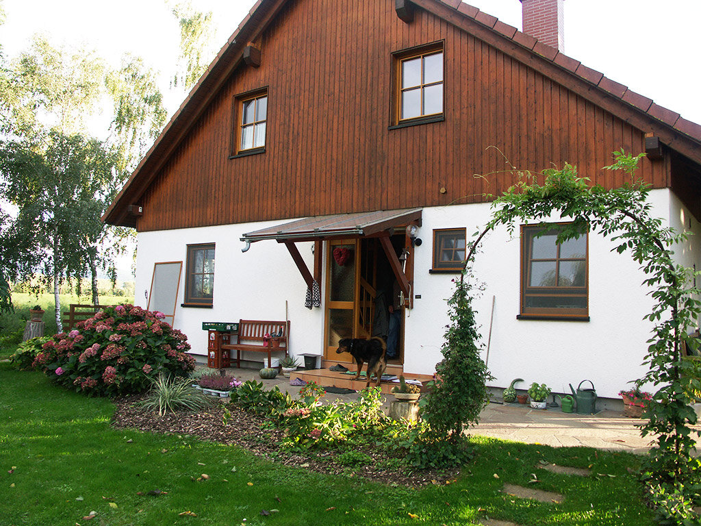 Einfamilienhaus mit weißer Putz-Fassade und braunem Holzgiebel; über der Haustür ein Vordach zur Wetterabsicherung, umgeben von einem gepflegten Garten mit Büschen und Rasen.