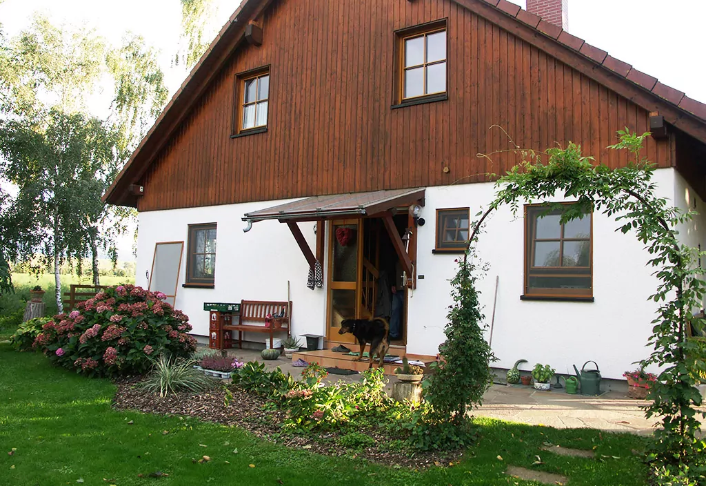 Stilvolle Terrasse mit Außenjalousien Einfamilienhaus mit weißer Putz-Fassade und braunem Holzgiebel; über der Haustür ein Vordach zur Wetterabsicherung, umgeben von einem gepflegten Garten mit Büschen und Rasen